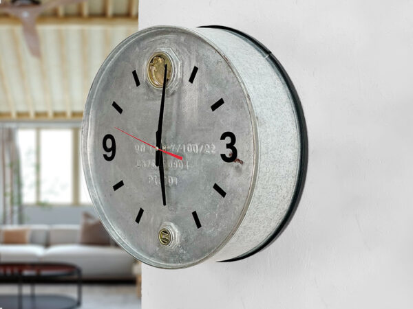 Verzinkte Ölfass Wanduhr mit Fassdeckeln auf der 6 und 12,schwarzem Ziffernblatt an einer weißen Wand, links verschwommen im Hintergrund ein graues Sofa mit schwarzem Tisch